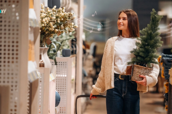**Alt text:** Smiling shopper carrying a small Christmas tree in a retail store with background music playing from shelf speakers